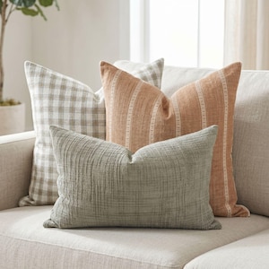 May include: Four decorative throw pillows in neutral tones rest on a beige sofa. The pillows feature various textures and patterns, including a gingham check, vertical stripes, and a woven design. The color palette includes shades of beige, tan, and sage green.