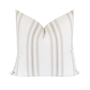 May include: A square decorative pillow with a white background and vertical beige stripes. The pillow has a soft, textured appearance and is likely made of linen or a similar fabric. The pillow is a neutral color and would complement a variety of home decor styles.