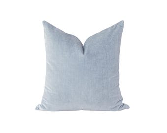 May include: A square throw pillow in a solid light blue color. The pillow features a textured fabric surface with a visible weave pattern and a soft, slightly plush appearance. The top center of the pillow has a distinct indentation or crease.