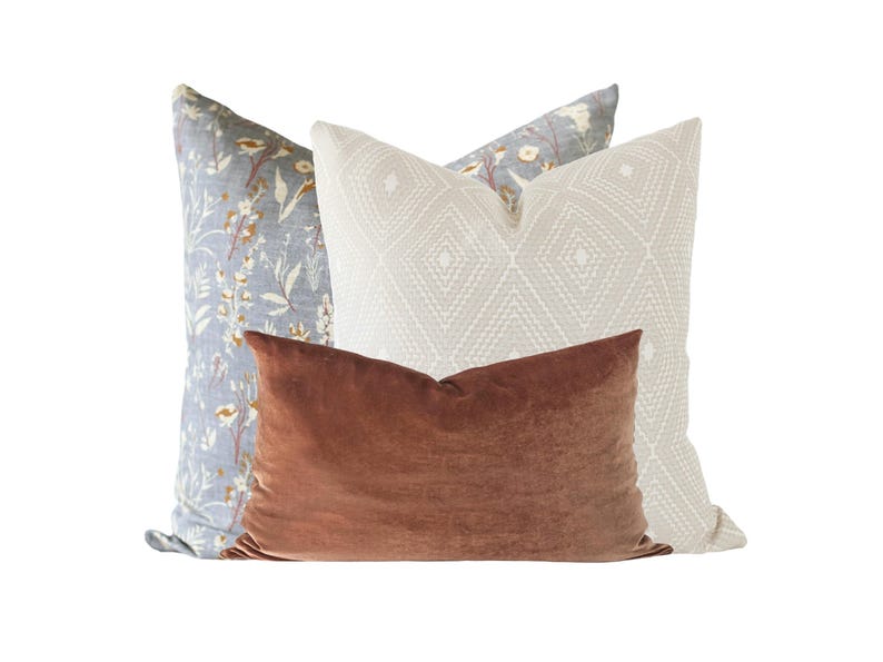 May include: Three decorative throw pillows. The front pillow is a rectangular, rust-colored velvet. Behind it is a white pillow with a diamond pattern, and a blue floral pillow.