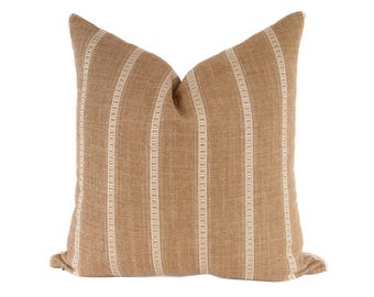 May include: A square throw cushion featuring a textured tan fabric with vertical stripes in a lighter cream colour. The stripes have a repeating geometric pattern within them, and the cushion has a soft, plump appearance with a visible seam along the edge.