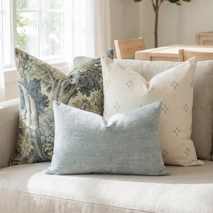 May include: A collection of decorative throw pillows in various patterns and colors on a neutral-toned sofa. Pillows include a light blue textured pillow, a cream-colored pillow with a small diamond pattern, and a pillow with a blue and green tree design.