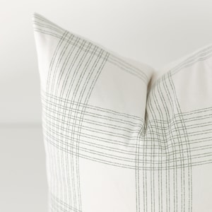 May include: A square throw pillow featuring a white fabric base with a pattern of thin, dark green lines arranged in a grid. The lines are composed of small, dashed segments that intersect to form a plaid design across the surface of the pillow.