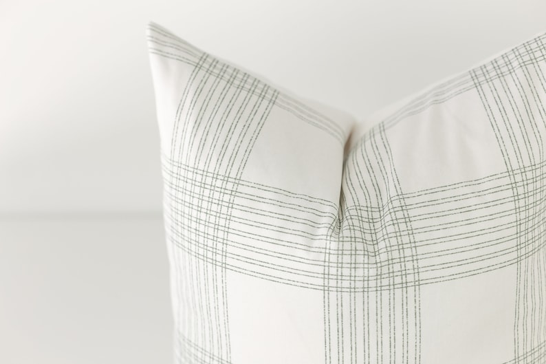 May include: A square throw pillow featuring a white fabric base with a pattern of thin, dark green lines arranged in a grid. The lines are composed of small, dashed segments that intersect to form a plaid design across the surface of the pillow.