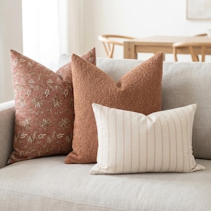 May include: Four decorative throw pillows on a light-colored sofa. The pillows feature various textures and patterns, including floral, textured knit, and striped designs. Colors include terracotta, cream, and beige, creating a warm and inviting aesthetic.