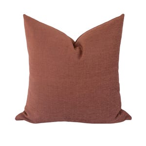 May include: A square throw pillow in a solid, muted terracotta or rust-colored fabric. The material features a subtle, fine-grained texture throughout the surface. The pillow is plumped at the top center, creating a slight indentation and soft folds.