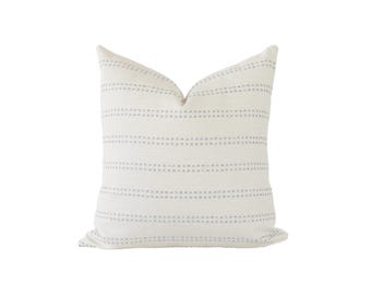 May include: A square throw pillow in an off-white color featuring horizontal rows of small, dashed gray lines. The fabric has a visible woven texture, and the pillow is shaped with a slight indentation at the top center edge.