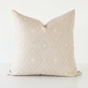 May include: A square beige pillow with a geometric diamond pattern. The pillow has a textured appearance and a neutral color palette, suitable for various home decor styles. The pillow is approximately 20 inches square.