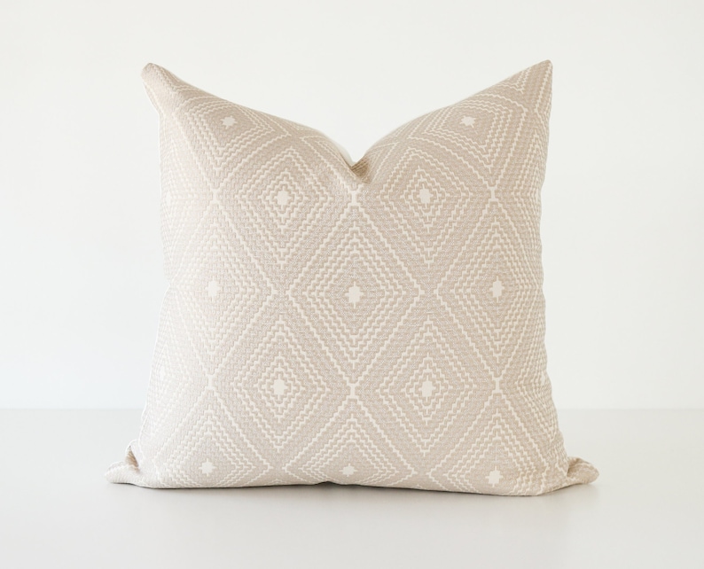 May include: A square beige pillow with a geometric diamond pattern. The pillow has a textured appearance and a neutral color palette, suitable for various home decor styles. The pillow is approximately 20 inches square.