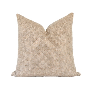 Puede incluir: Un cojín cuadrado de color beige claro con un patrón geométrico repetitivo de diamantes en toda la superficie. La tela tiene una apariencia tejida y texturizada con una profundidad sutil creada por las líneas en relieve del diseño.