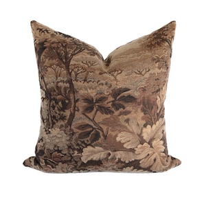 May include: A decorative throw pillow with a forest scene design. The pillow features a tapestry-like pattern in shades of brown, tan, and beige, depicting trees, foliage, and a landscape. The pillow is square-shaped and has a soft, textured appearance.
