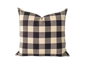 May include: A square throw pillow featuring a black and off-white buffalo check pattern. The fabric has a visible woven texture, and the pillow is filled to create a plump, structured shape with defined corners and a soft, rounded top edge.