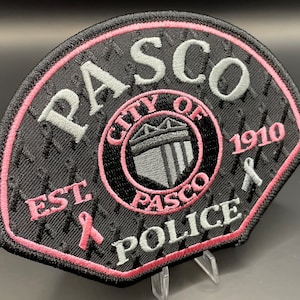 May include: Black and white embroidered patch with pink trim. The patch features the words "PASCO CITY OF PASCO 1910 POLICE" and a stylized shield with a crown. There are pink ribbons on either side of the patch.