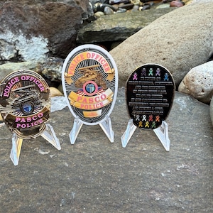 Może przedstawiać: Trzy pamiątkowe monety Pasco Police na przezroczystych stojakach. Monety mają złote i różowe akcenty, z napisami "Police Officer" i "Pasco Police". Jedna moneta zawiera przesłanie pamiątkowe i kolorowe wstążki.