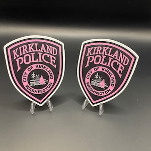 May include: Two black and white Kirkland Police Department patches with pink lettering and a sailboat graphic. The patches are on clear plastic stands.