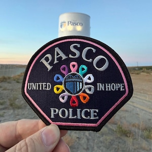 May include: A black embroidered patch with a pink border featuring the text PASCO UNITED IN HOPE POLICE. The center displays a blue shield icon surrounded by a circular arrangement of multi-colored awareness ribbons in various bright hues.