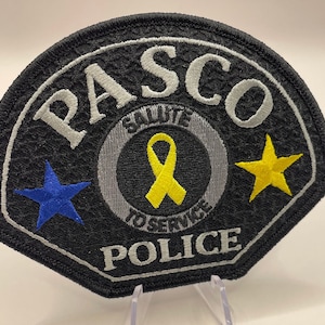 May include: Embroidered black police patch with the word "PASCO" in white, a yellow ribbon, and the words "SALUTE TO SERVICE POLICE". Features a blue and a yellow star.