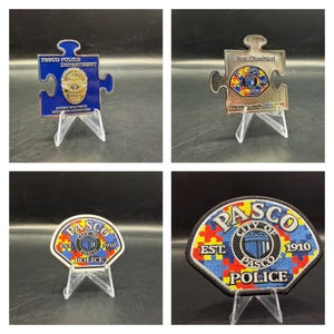 May include: Four different versions of a police badge with a puzzle piece design. The badges are blue, silver, and gray with a yellow, red, and blue puzzle piece design. The badges have the words "Pasco Police Department" and "City of Pasco" on them. The badges are on display stands.