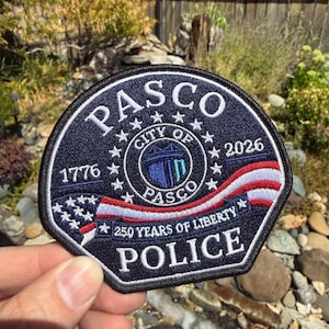 May include: A circular embroidered patch featuring a dark blue background with the text PASCO at the top and POLICE at the bottom. The center displays the City of Pasco seal, the years 1776 and 2026, and a waving American flag with 250 YEARS OF LIBERTY.