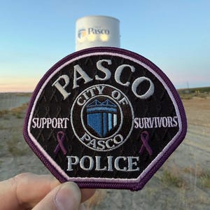 Könnte beinhalten: Ein kreisförmiger bestickter Aufnäher mit dunkelviolettem Rand und schwarzem Hintergrund. Er zeigt den Text PASCO, CITY OF PASCO, SUPPORT, SURVIVORS und POLICE in Weiß. Ein zentrales blaues Schildelement wird von zwei kleinen violetten Schleifen flankiert.