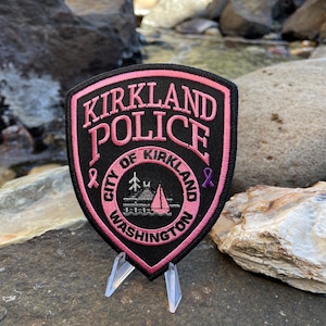 May include: Pink and black embroidered patch with the text "Kirkland Police" and "City of Kirkland Washington". The patch features a small sailboat and a pink ribbon.