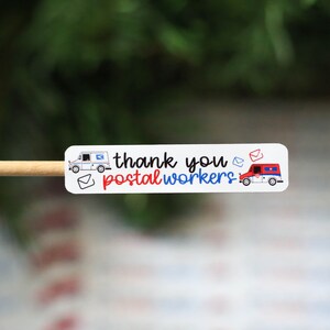 May include: A white sticker with the text "thank you postal workers" in black, red, and blue. The sticker features illustrations of mail trucks and envelopes. The sticker is on a wooden stick.
