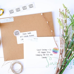 May include: A still-life composition featuring stationery and decorative elements. Includes brown envelopes, white address labels, a roll of twine, scissors, washi tape, and floral accents. The labels read "Happy Mail."