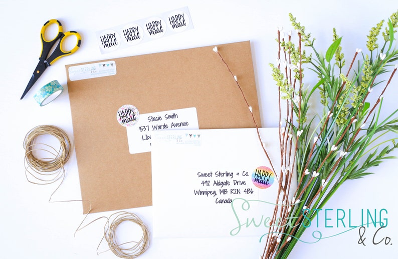 May include: A still-life composition featuring stationery and decorative elements. Includes brown envelopes, white address labels, a roll of twine, scissors, washi tape, and floral accents. The labels read "Happy Mail."