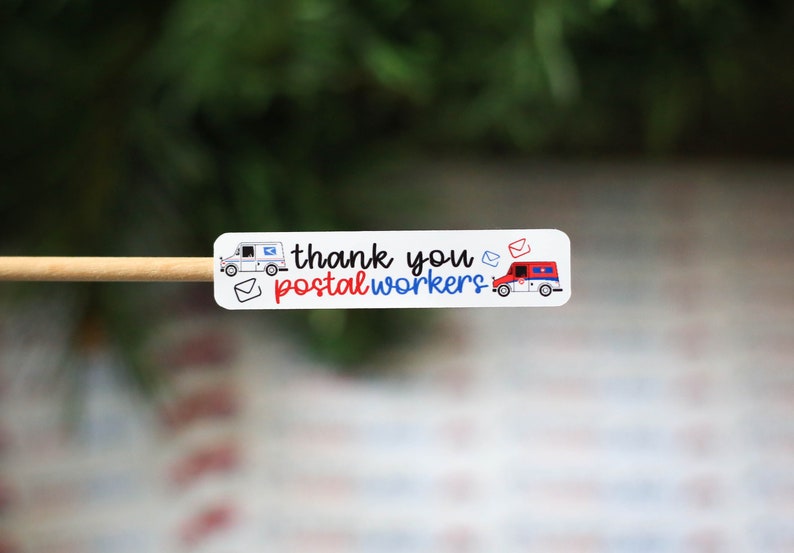May include: A white sticker with the text "thank you postal workers" in black, red, and blue. The sticker features illustrations of mail trucks and envelopes. The sticker is on a wooden stick.