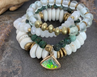 May include: A stack of four bracelets featuring white, green, and gold beads. The bottom bracelet has a gold charm with an iridescent stone.