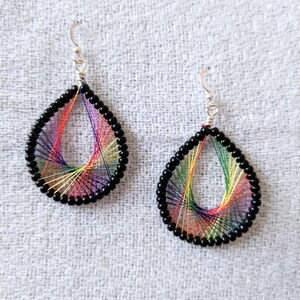 May include: A pair of teardrop earrings. Each earring has a rainbow of threads woven within a black beaded frame. The earrings have silver ear wires and are displayed on a light grey textured surface.