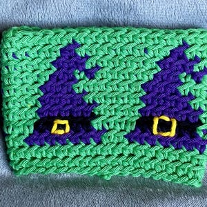 May include: A rectangular crochet cup cozy featuring a bright green background with two purple witch hats. Each hat has a black band and a yellow square buckle. The texture is created with visible yarn stitches in a consistent, repeating pattern.