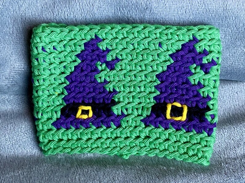 May include: A rectangular crochet cup cozy featuring a bright green background with two purple witch hats. Each hat has a black band and a yellow square buckle. The texture is created with visible yarn stitches in a consistent, repeating pattern.