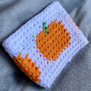 May include: A rectangular crochet pouch featuring a white background with an orange pumpkin design in the center. The pumpkin has a small green stem at the top. The crochet work is constructed with a dense, textured stitch pattern throughout the item.