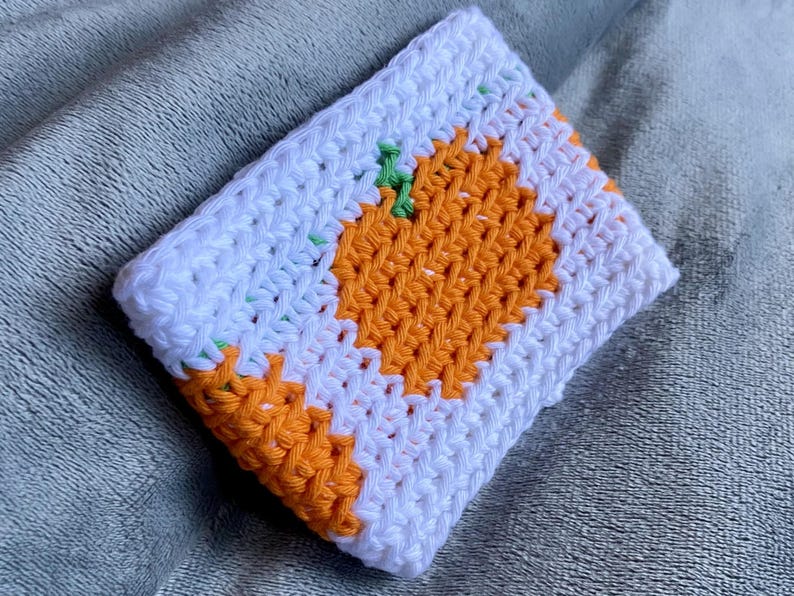 May include: A rectangular crochet pouch featuring a white background with an orange pumpkin design in the center. The pumpkin has a small green stem at the top. The crochet work is constructed with a dense, textured stitch pattern throughout the item.