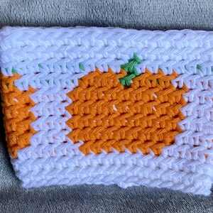May include: A rectangular crochet cup sleeve featuring a white background with a repeating orange pumpkin pattern. The pumpkins are rendered in orange yarn with small green stems, and the entire piece is constructed with a consistent stitch.