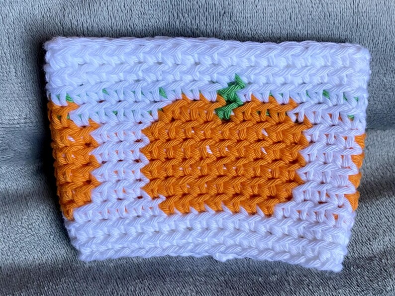May include: A rectangular crochet cup sleeve featuring a white background with a repeating orange pumpkin pattern. The pumpkins are rendered in orange yarn with small green stems, and the entire piece is constructed with a consistent stitch.