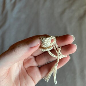 A crocheted teacup bookmark held by hand in front of a gray cloth background. The teacup is held with the thumb and index fingers and the string is wrapped around the ring and middle fingers.