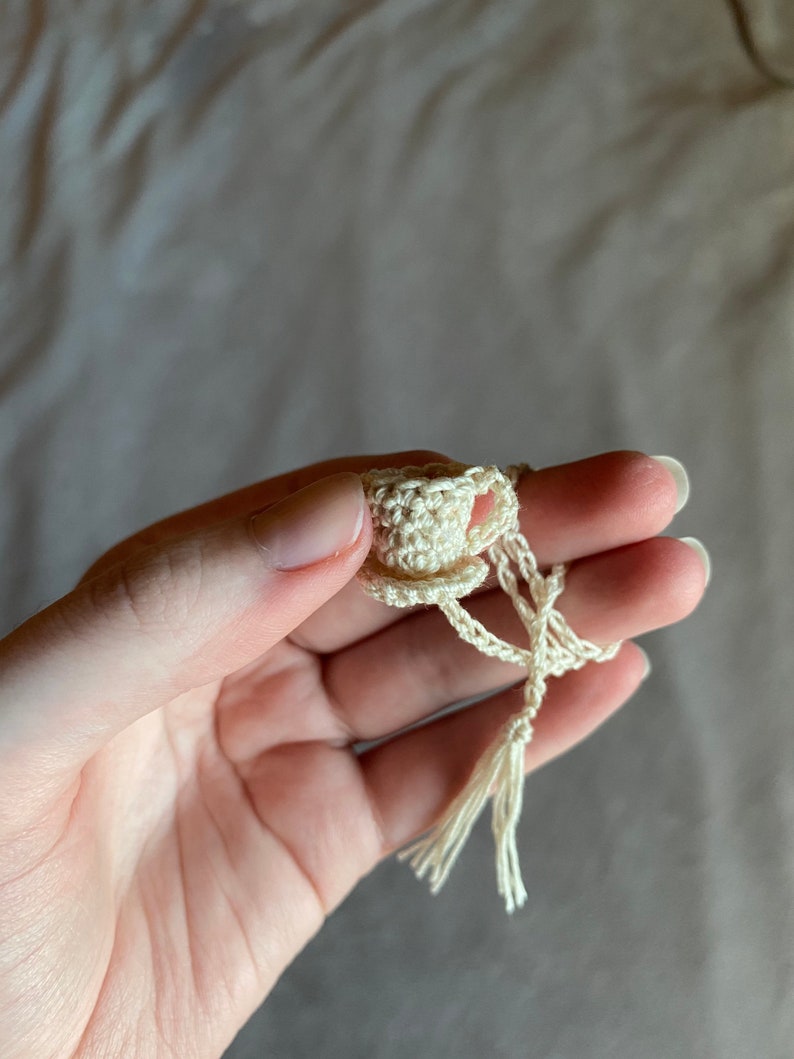 A crocheted teacup bookmark held by hand in front of a gray cloth background. The teacup is held with the thumb and index fingers and the string is wrapped around the ring and middle fingers.