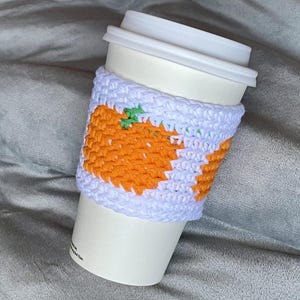 May include: A white disposable coffee cup featuring a handmade crochet sleeve. The sleeve is crafted from light lavender yarn and displays a repeating pattern of bright orange pumpkins with small green stems, providing a textured grip.