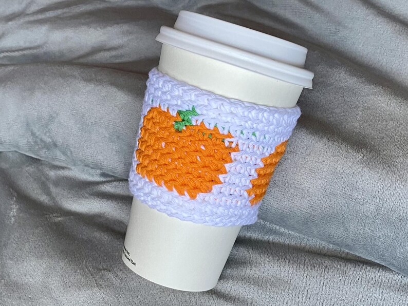 May include: A white disposable coffee cup featuring a handmade crochet sleeve. The sleeve is crafted from light lavender yarn and displays a repeating pattern of bright orange pumpkins with small green stems, providing a textured grip.