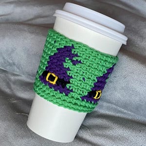 May include: A white disposable coffee cup featuring a green crocheted sleeve. The sleeve displays a repeating pattern of purple witch hats with black bands and yellow buckles, crafted with a textured knit stitch throughout the design.