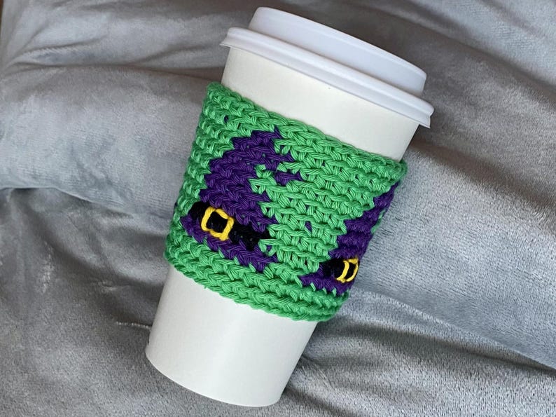 May include: A white disposable coffee cup featuring a green crocheted sleeve. The sleeve displays a repeating pattern of purple witch hats with black bands and yellow buckles, crafted with a textured knit stitch throughout the design.