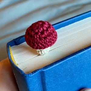 May include: A small, handmade crochet bookmark shaped like a mushroom with a deep red cap and a cream-colored stem. The mushroom is positioned between the pages of a closed blue hardcover book, with the cap resting on the top edge of the pages.