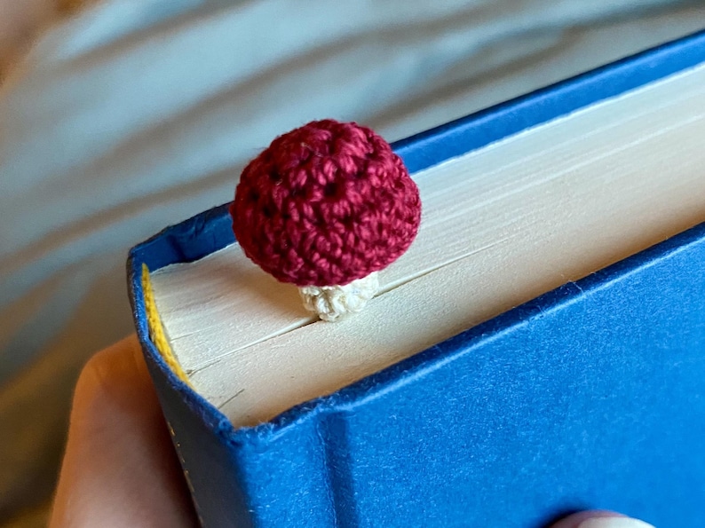May include: A small, handmade crochet bookmark shaped like a mushroom with a deep red cap and a cream-colored stem. The mushroom is positioned between the pages of a closed blue hardcover book, with the cap resting on the top edge of the pages.