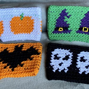 May include: Four rectangular crochet cup sleeves featuring seasonal designs. One white sleeve with orange pumpkins, one green sleeve with purple witch hats, one orange sleeve with a black bat, and one black sleeve with two white skulls.