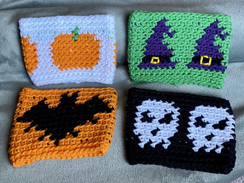 May include: Four rectangular crochet cup sleeves featuring seasonal designs. One white sleeve with orange pumpkins, one green sleeve with purple witch hats, one orange sleeve with a black bat, and one black sleeve with two white skulls.