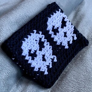 May include: A rectangular crochet pouch featuring a black background with two white skull motifs. The item is constructed with a dense, textured stitch pattern, showcasing a high-contrast design with clear, defined edges for the skull shapes.