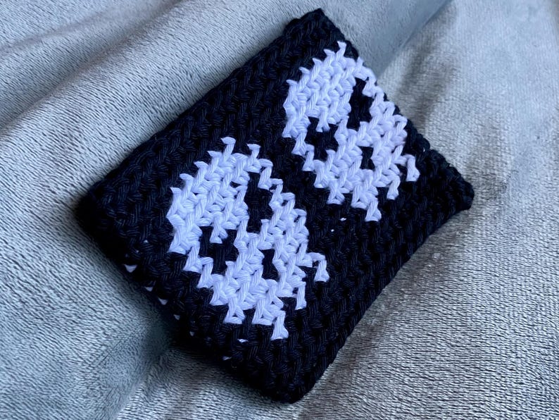 May include: A rectangular crochet pouch featuring a black background with two white skull motifs. The item is constructed with a dense, textured stitch pattern, showcasing a high-contrast design with clear, defined edges for the skull shapes.