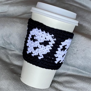 May include: A white disposable coffee cup with a white plastic lid, featuring a black and white crocheted sleeve. The sleeve displays a repeating pattern of white ghost shapes against a solid black background, wrapped around the center of the cup.